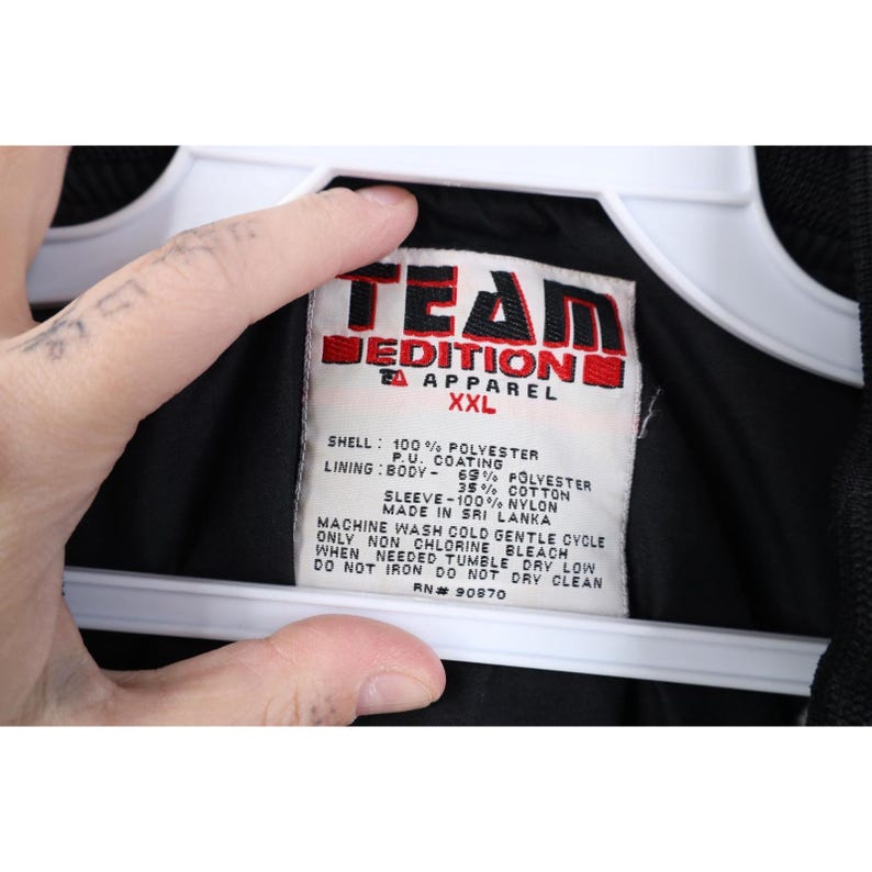 May include: A black Team Edition Apparel XXL jacket label. The label is white with red and black text, listing the shell and lining materials. The jacket is made in Sri Lanka.