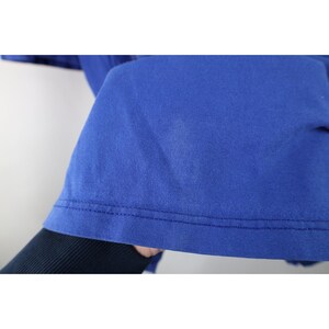May include: Close-up of a royal blue t-shirt. The fabric is smooth and appears to be cotton. The hem is visible, with a double-stitched seam. The shirt is held up, revealing a glimpse of a dark sleeve underneath.