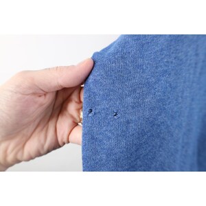 Op de afbeelding: Een kledingstuk van blauwe gem&ecirc;leerde gebreide stof met twee kleine, duidelijke gaatjes bij de rand van het materiaal. De textuur van de stof is zichtbaar en toont een consistent weefpatroon over het gehele blauwe textieloppervlak.