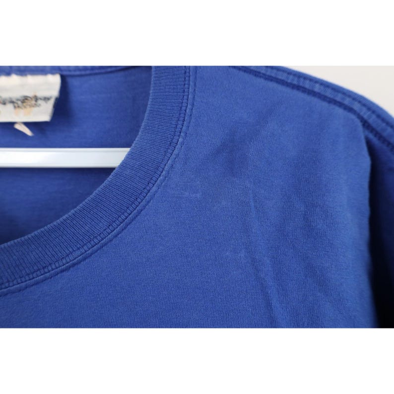 May include: Close-up of a royal blue t-shirt. The shirt has a crew neck and short sleeves. A small tag is visible on the upper left side. The fabric appears to be soft and slightly wrinkled.