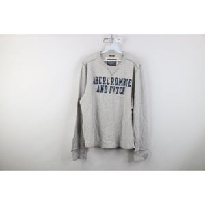May include: A light grey Abercrombie and Fitch long-sleeved sweatshirt with a crew neck. The brand name is printed in navy blue across the chest. The sweatshirt is hanging on a white hanger.