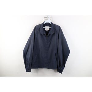 May include: A navy blue zip-up jacket featuring a pointed collar, long sleeves with buttoned cuffs, and two front pockets. The garment includes a central zipper closure and a white interior brand label visible at the neckline, hanging on a hanger.