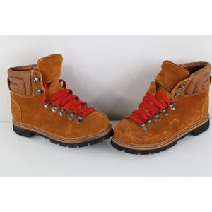 May include: A pair of tan suede hiking boots featuring bright red laces, silver-toned metal eyelets, and a padded brown collar. The boots have a thick black rubber lug sole and a rounded toe design, providing a sturdy look for outdoor activities.