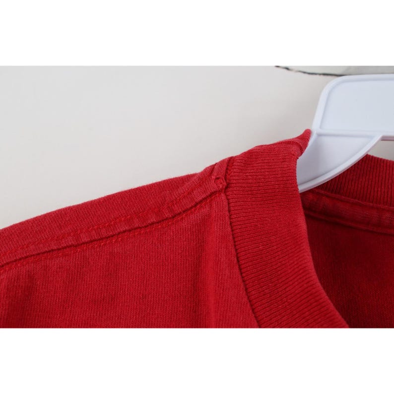 May include: Close-up of a red t-shirt, showing the shoulder and neckline. The fabric is a solid, vibrant red, with visible stitching along the seams. The shirt is hanging on a white hanger.