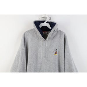May include: A heather gray long-sleeve hooded sweatshirt featuring a small embroidered Mickey Mouse character on the left chest. The hoodie includes a dark navy blue inner lining, a matching drawstring, and a white plastic hanger at the neck.