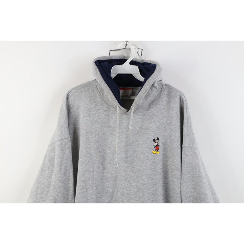 May include: A heather gray long-sleeve hooded sweatshirt featuring a small embroidered Mickey Mouse character on the left chest. The hoodie includes a dark navy blue inner lining, a matching drawstring, and a white plastic hanger at the neck.