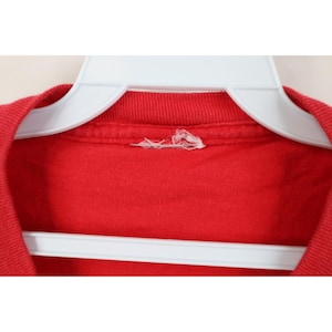 May include: Close-up of a red t-shirt hanging on a white plastic hanger. The shirt's collar and shoulder area are visible, with a small tear near the tag. The fabric appears to be a solid, vibrant red.