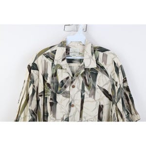 Op de afbeelding: Een overhemd met korte mouwen en een bamboe-print in groen, grijs en beige. Het overhemd heeft een kraag en hangt aan een witte hanger. Het overhemd is bedrukt met bamboebladeren.