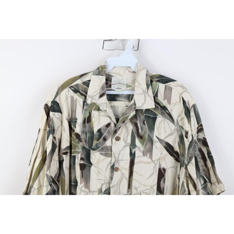 Op de afbeelding: Een overhemd met korte mouwen en een bamboe-print in groen, grijs en beige. Het overhemd heeft een kraag en hangt aan een witte hanger. Het overhemd is bedrukt met bamboebladeren.