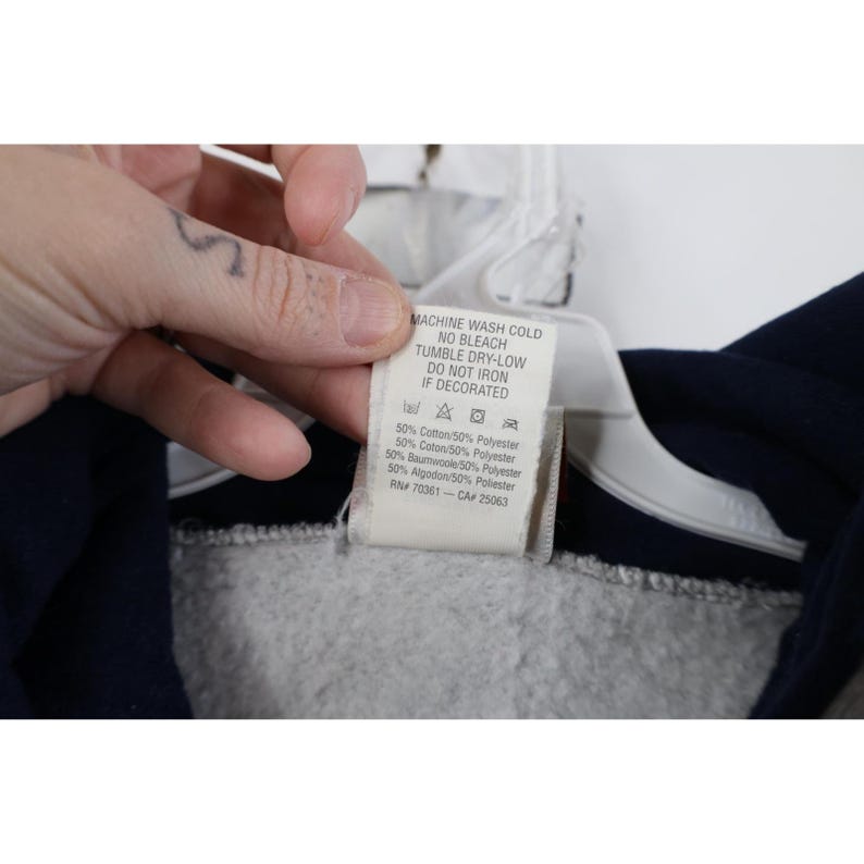 May include: A white fabric garment care tag attached to a dark blue and grey sweatshirt. The tag features black text including MACHINE WASH COLD, NO BLEACH, TUMBLE DRY-LOW, DO NOT IRON IF DECORATED, and material composition of 50% Cotton/50% Polyester.