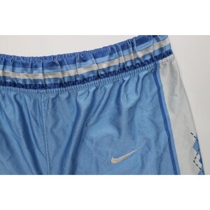 May include: Light blue athletic shorts featuring a silver Nike swoosh logo on the front. The waistband is elasticized with horizontal stripes in shades of blue and white. A vertical white panel runs down the side with a dark blue diamond pattern.