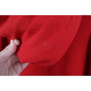 Peut inclure: Gros plan sur un tissu rouge vif, possiblement un v&ecirc;tement. La texture du mat&eacute;riau est visible, avec de l&eacute;g&egrave;res variations dans le tissage. Le tissu est tenu, avec une main visible dans le cadre.