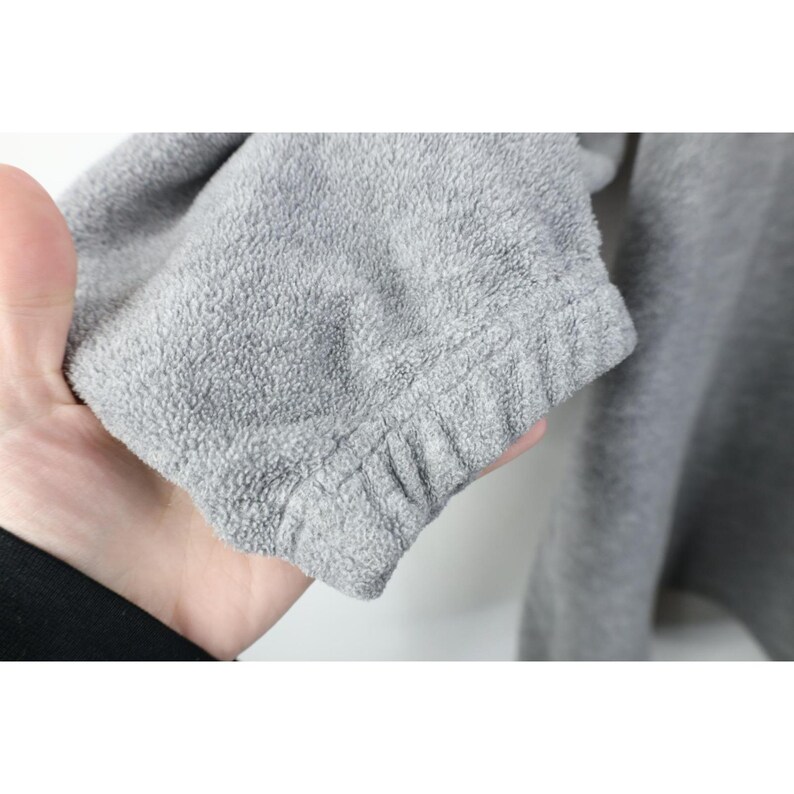 May include: Close-up of a gray fleece fabric sleeve. The sleeve has a soft, textured appearance and a ribbed cuff. The fabric appears to be a light gray color. The sleeve is part of a larger garment.