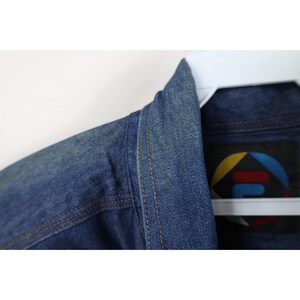 May include: Close-up of a blue denim jacket collar and shoulder. The jacket has visible stitching in a contrasting tan color. A label with a colorful geometric design is visible.