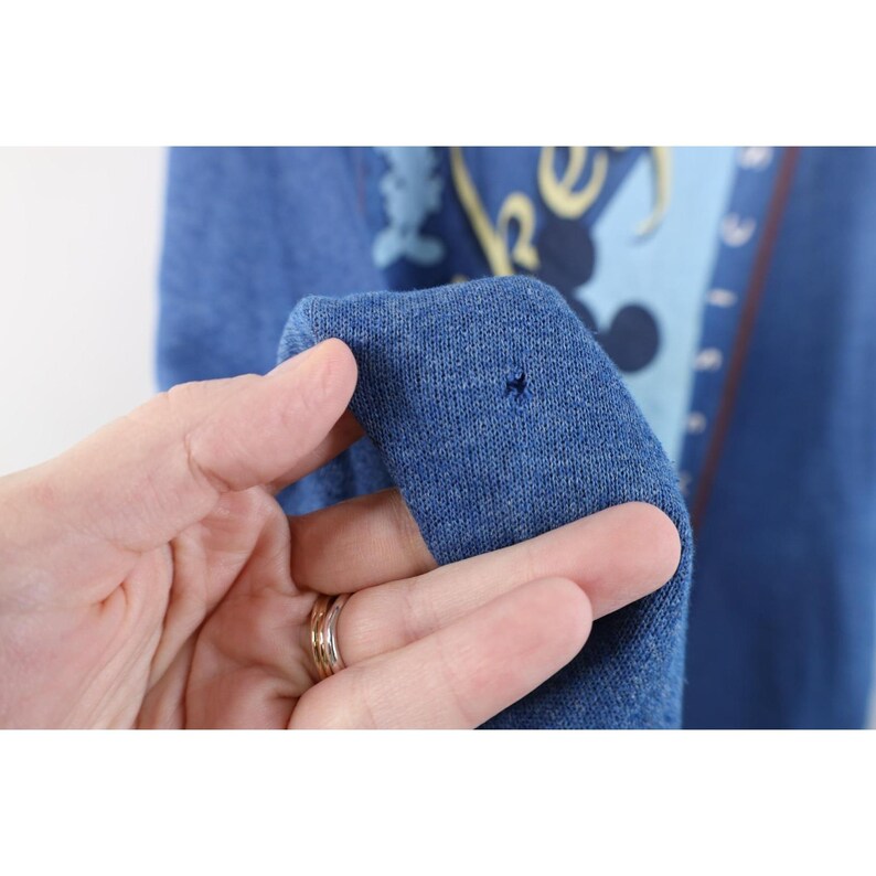 Op de afbeelding: Een blauwe gem&ecirc;leerde sweater met een grafische print met het woord Classics en een donkerblauw silhouet van een muisfiguur. De stof vertoont een klein gaatje bij het midden, waardoor de textuur van het gebreide materiaal zichtbaar is.