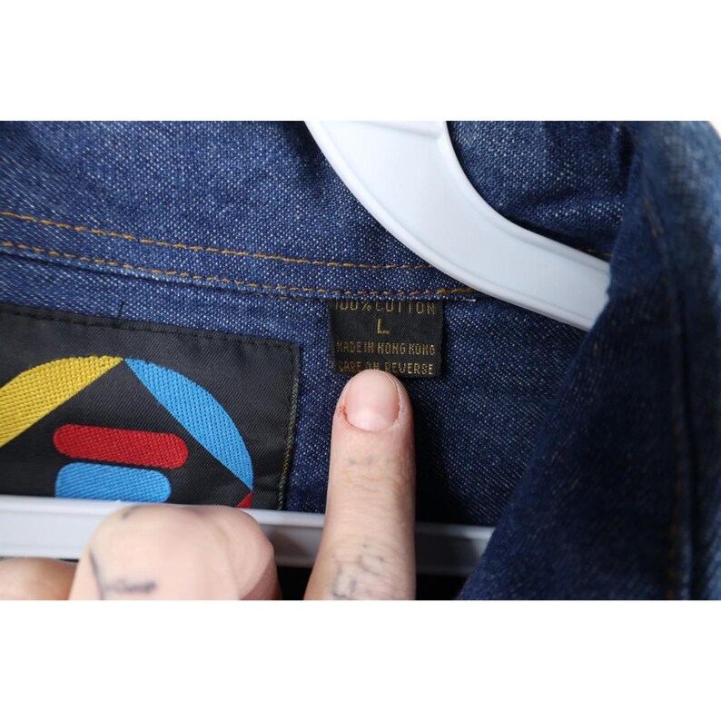 May include: Close-up of a blue denim jacket with a tag that reads "100% Cotton" and "Made in Hong Kong". The tag also features a colorful logo. The jacket is hanging on a white hanger.