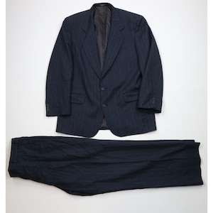Puede incluir: Un traje de color azul marino oscuro que consta de una americana de dos botones con solapas de muesca y pantalones a juego. La tela presenta un sutil patrón de rayas verticales. La americana incluye dos bolsillos con solapa y tres botones.