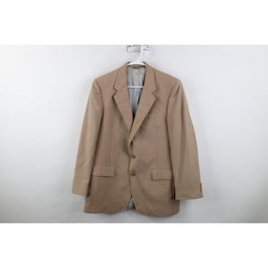May include: A tan blazer featuring a notched lapel, a two-button front closure, and two flap pockets at the waist. The garment has long sleeves with buttoned cuffs and a visible light-coloured interior lining visible at the collar area.