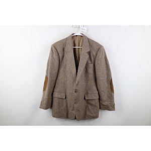 May include: A brown tweed blazer with two front pockets and brown suede elbow patches. The blazer is buttoned and hanging on a white hanger against a white background. The blazer has a notched lapel.