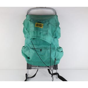 May include: A teal external frame backpack featuring a silver metal frame, black adjustable straps, and a rectangular yellow patch with the black text ROTHCO centered on the top flap. The bag includes multiple side pockets and a front zippered compartment.