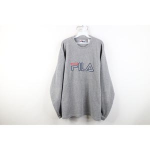 May include: A gray long-sleeve sweatshirt with the brand name "FILA" in red and navy blue lettering. The sweatshirt is hanging on a white hanger against a white background. The brand name is centered on the chest.