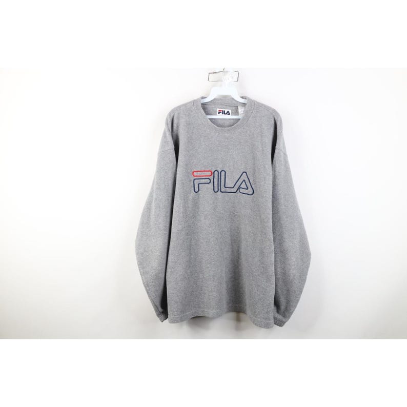 May include: A gray long-sleeve sweatshirt with the brand name "FILA" in red and navy blue lettering. The sweatshirt is hanging on a white hanger against a white background. The brand name is centered on the chest.