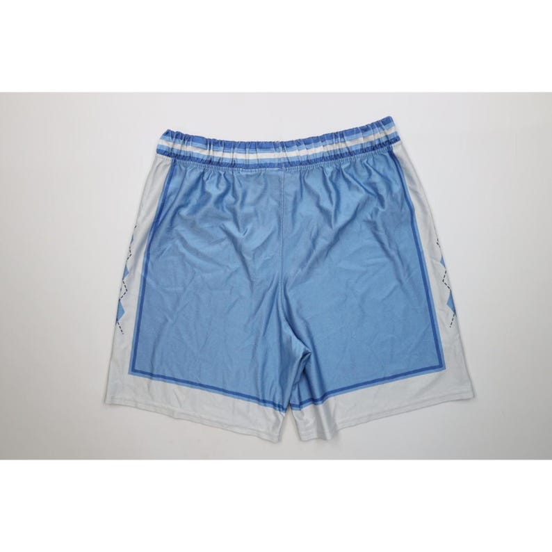 May include: Light blue athletic shorts featuring a white waistband with blue stripes, a solid light blue center panel framed by a darker blue border, and white side panels decorated with a black and blue diamond argyle pattern.