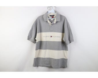 Könnte beinhalten: Ein grau-weiß gestreiftes Poloshirt mit kurzen Ärmeln. Das Shirt hat einen weißen horizontalen Streifen über der Brust mit dem Text "TOMMY HILFIGER" in dunkler Schrift. Es hat einen Kragen und eine Knopfleiste mit drei Knöpfen.