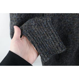 May include: Close-up of a textured, dark gray sweater sleeve with a ribbed cuff. The cuff features a blend of blue, brown, and gray yarns. The sweater appears to be made of a warm, possibly wool, material.