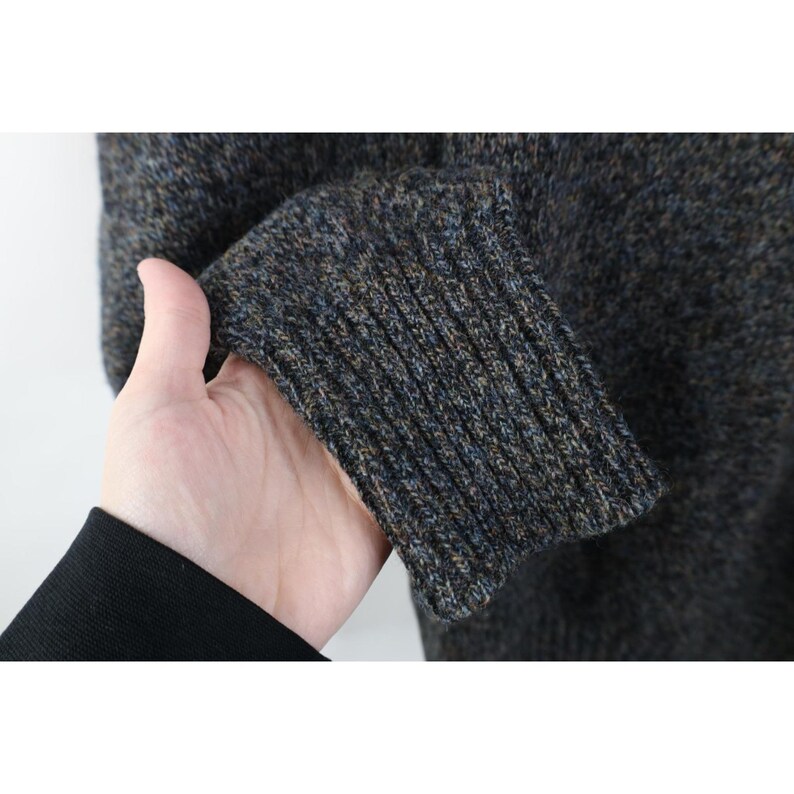 May include: Close-up of a textured, dark gray sweater sleeve with a ribbed cuff. The cuff features a blend of blue, brown, and gray yarns. The sweater appears to be made of a warm, possibly wool, material.