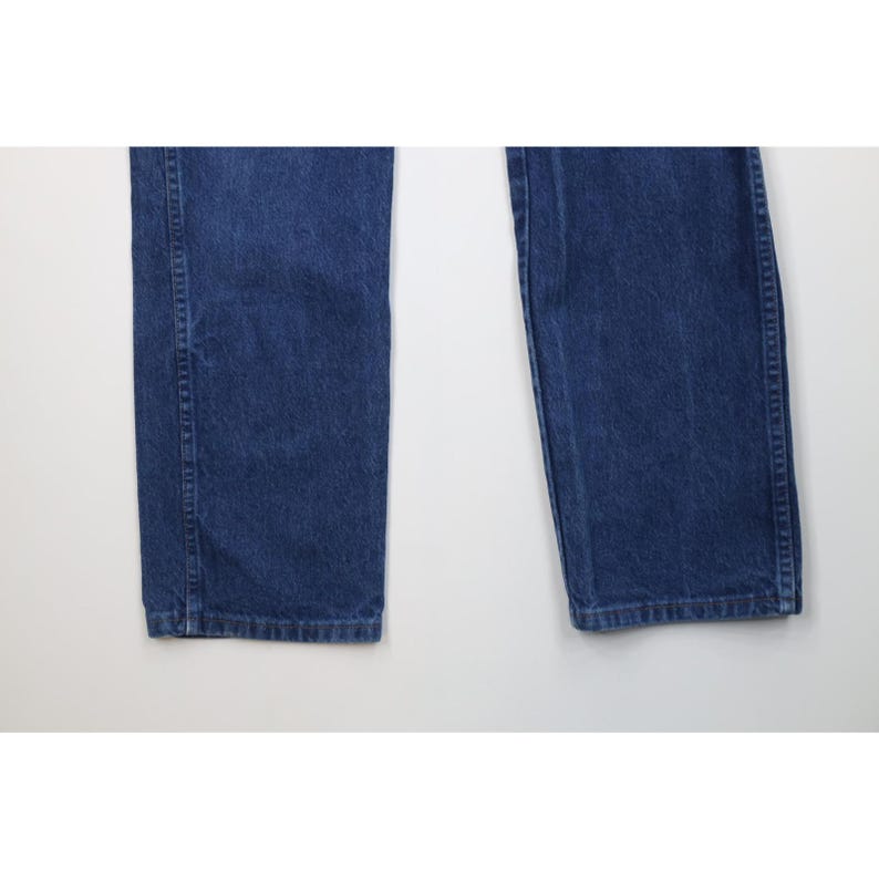 May include: Close-up of the lower portion of a pair of blue denim jeans. The jeans are a medium wash and have a classic straight-leg fit. The image shows the legs of the jeans from the knees down, with the hems visible.