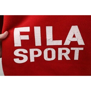 May include: Close-up of a red fabric with the white text "FILA SPORT". The letters have a distressed, cracked appearance. The fabric is held by a hand on the left side of the image. The overall theme is athletic apparel.