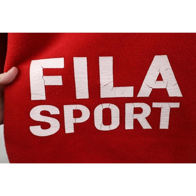 May include: Close-up of a red fabric with the white text "FILA SPORT". The letters have a distressed, cracked appearance. The fabric is held by a hand on the left side of the image. The overall theme is athletic apparel.