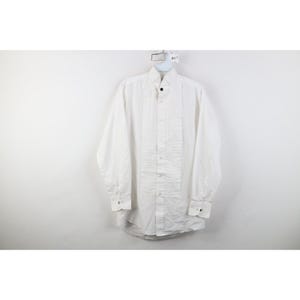 Pode incluir: Uma camisa branca com frente de peito plissada e mangas compridas. A camisa tem uma gola clássica e punhos abotoados. Está pendurada num cabide branco sobre um fundo branco.