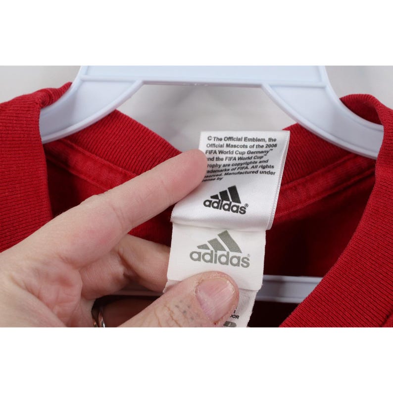 May include: Close-up of a red t-shirt with a white tag featuring the Adidas logo and text. The tag also includes text about the 2006 FIFA World Cup Germany.