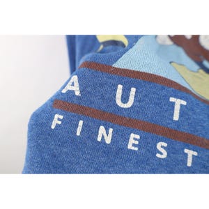 Op de afbeelding: Een blauw shirt met lange mouwen voorzien van een grafische print met de tekst AUT en FINEST in witte hoofdletters. Het ontwerp bevat horizontale bruine strepen en een lichtblauwe en gele illustratie op een textuurrijke blauwe stof.
