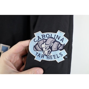 May include: Black jacket with a Carolina Tar Heels patch on the sleeve. The patch features a light blue and white design with the words "Carolina Tar Heels" surrounding a ram graphic.