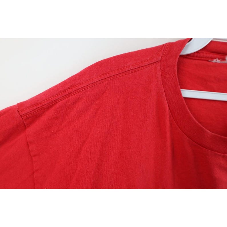 May include: Close-up of a solid red t-shirt. The image focuses on the shoulder and neckline, showing the stitching details. The shirt is hanging on a white hanger against a plain white background. The fabric appears to be cotton.
