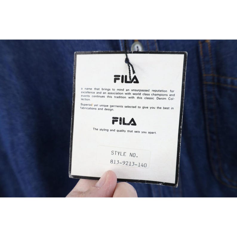 May include: A close-up of a Fila brand tag attached to a denim garment. The tag features the Fila logo and text describing the brand's reputation and quality. The style number 813-9213-140 is visible.