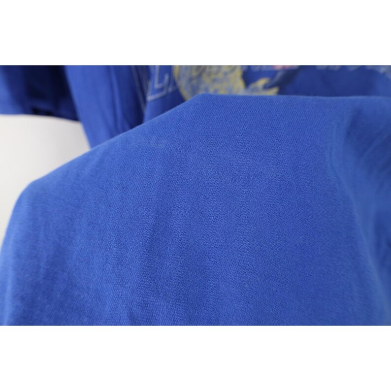 May include: Close-up of a royal blue t-shirt. The fabric is soft and slightly wrinkled, with a subtle texture. The image focuses on the shirt's material and color, highlighting its solid hue and the way the light interacts with the fabric.
