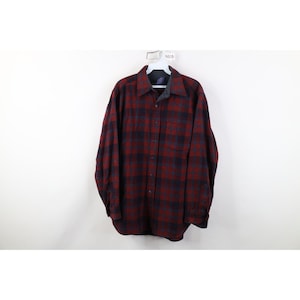 May include: A long-sleeved, button-down flannel shirt in a red and navy blue plaid pattern. The shirt is hanging on a white hanger against a white background. The shirt has a collared neckline and a single chest pocket.