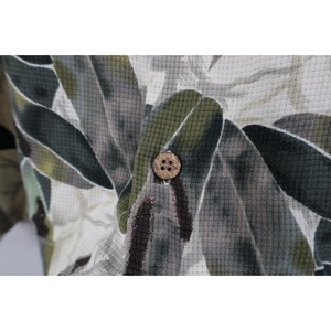 Op de afbeelding: Close-up van een overhemd met knopen met een bladerenpatroon in groen, grijs en wit. Het overhemd heeft een textuurstof en een natuurlijk gekleurde knoop.