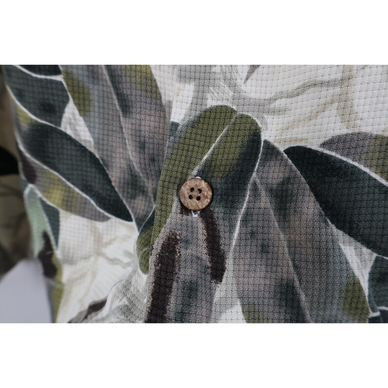 Op de afbeelding: Close-up van een overhemd met knopen met een bladerenpatroon in groen, grijs en wit. Het overhemd heeft een textuurstof en een natuurlijk gekleurde knoop.