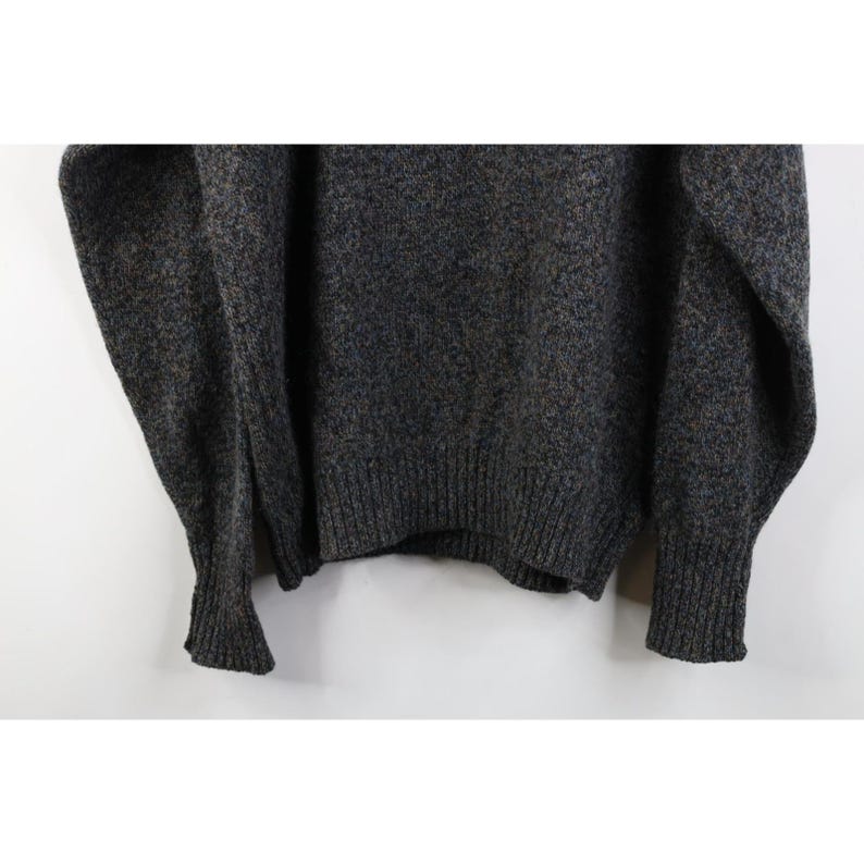 May include: A dark gray sweater with a ribbed hem and cuffs. The sweater has a speckled texture with hints of blue and brown. The sleeves are long and the overall style is casual and comfortable.