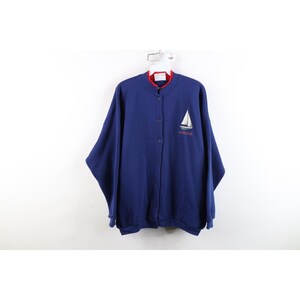 May include: A navy blue button-up sweatshirt featuring a mock neck collar with a red inner lining. The front displays an embroidered graphic of a white sailboat with the text "SAILOR PLAIN" underneath. The garment has long sleeves and ribbed cuffs.