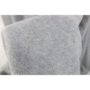 May include: Close-up of a light gray fleece fabric with a textured surface. The fabric has small brown spots scattered across the surface. The fleece is folded over, revealing the soft texture.