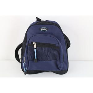 May include: A navy blue and black backpack featuring a front zippered pocket with a black mesh overlay. The bag includes a top handle, adjustable straps, and a small oval patch with the text Mudd in white letters on a blue background.