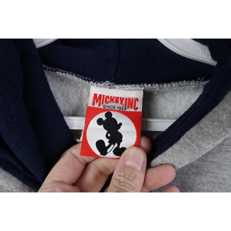 May include: A grey hooded sweatshirt with a dark navy blue hood lining. The interior neck label is white with a red rectangular border, featuring the black silhouette of Mickey Mouse and the red text Mickey Inc. Since 1928.