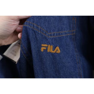 May include: Close-up of a dark blue denim jacket with the FILA logo embroidered in gold thread. The jacket features gold stitching and a tag is visible. The denim fabric has a classic, worn look.
