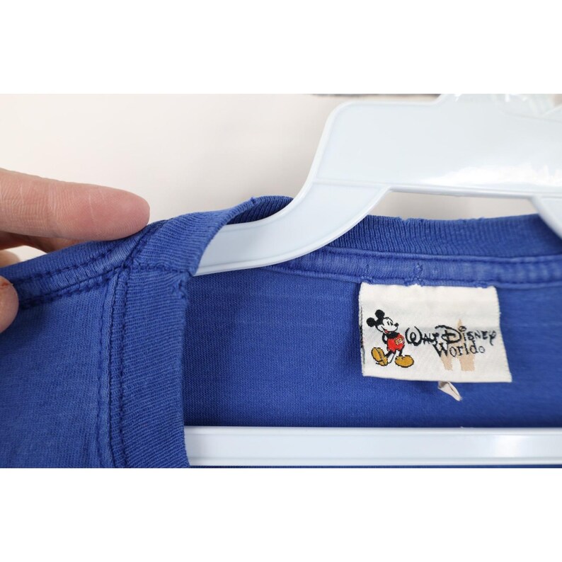 May include: Close-up of a blue t-shirt with a Walt Disney World tag. The tag features a graphic of Mickey Mouse and the words "Walt Disney World". The shirt is hanging on a white hanger.